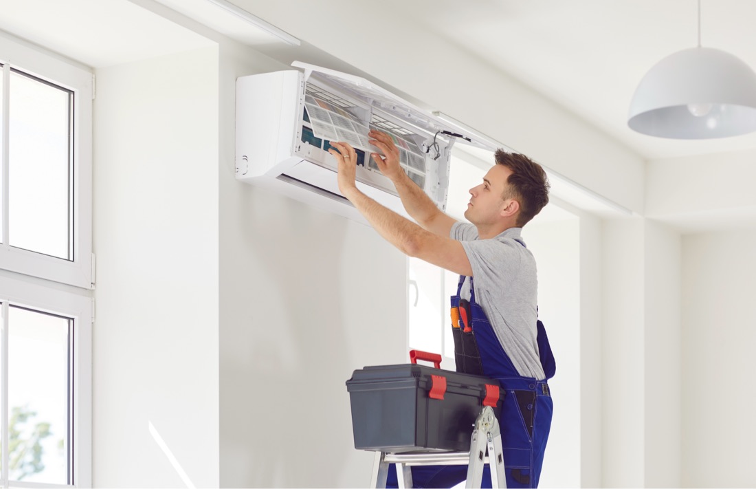 A technician on a ladder working on a mounted AC unit indoors